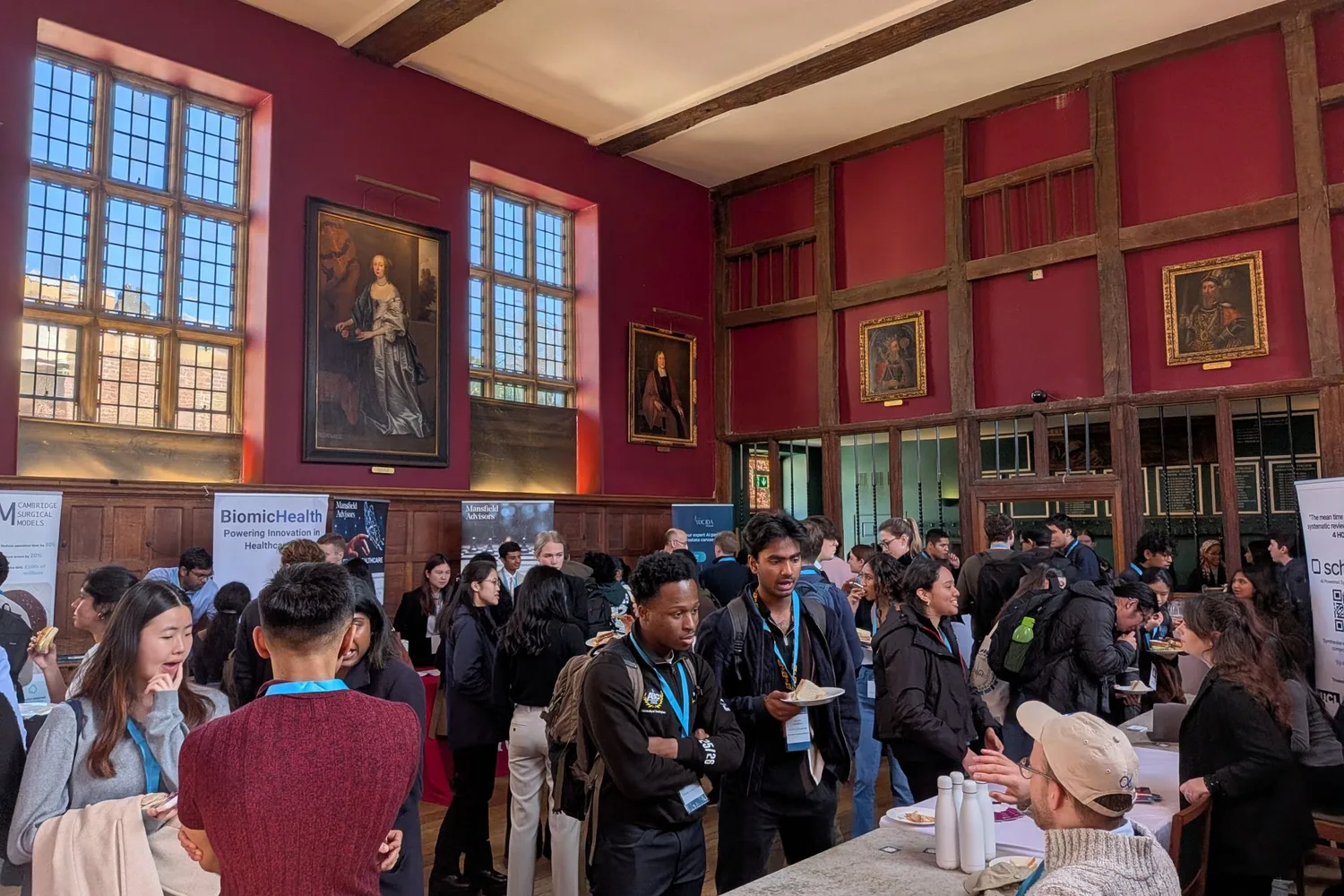 Delegates and exhibitors networking in a panelled hall with red walls and portraits at the Cambridge Health Futures Conference, with sponsor banners along the far wall